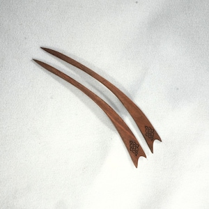 Könnte beinhalten: Zwei Haarstäbe aus Holz in dunkler Braunfärbung. Jeder Stab hat eine gebogene Form mit einer Spitze und einem keltischen Knotenmuster, das nahe der Basis eingraviert ist. Präsentiert auf weißem Hintergrund.