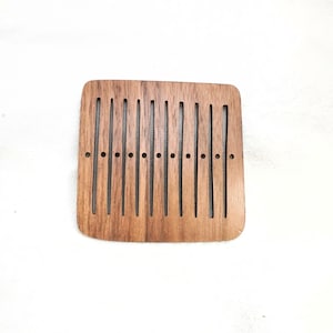 Pode incluir: Um pente de madeira retangular com ranhuras verticais e pequenos orifícios. O pente é feito de madeira castanha escura e tem um acabamento liso. Projetado para cuidados e penteados capilares, mede cerca de 10 cm de comprimento.