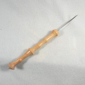 Könnte beinhalten: Ein Holzwerkzeug mit einer langen, dünnen Metallnadel. Der Griff ist aus hellem Holz mit einer natürlichen Oberfläche. Das Werkzeug wird zum Basteln und Nähen verwendet.