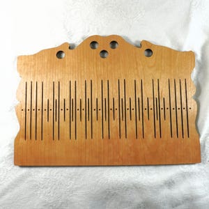 May include: A light-colored wooden weaving tool with a decorative top edge featuring three circular holes. The tool has vertical slots and small holes, likely for weaving. The wood grain is visible, and the tool is set against a white background.