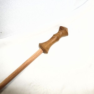 May include: A handcrafted wooden wand with a light brown shaft and a darker, intricately carved handle. The wand's handle has a unique shape, adding to its aesthetic appeal. The wand is made of wood and is a magical item.