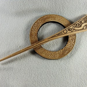 Könnte beinhalten: Eine Brosche aus Holz mit keltischen Knotenmustern und einem langen, dünnen Stift. Die Brosche ist rund mit komplizierten Knotenmustern und der Stift hat oben ein keltisches Knotenmuster.