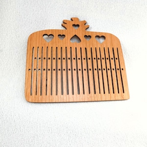 Puede incluir: Un peine de telar de madera con diseño de corazón. El peine es de madera marrón claro y presenta ranuras verticales y pequeños agujeros. La parte superior tiene tres recortes de corazón y una corona decorativa. Mide unos 20 cm.