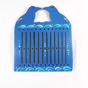 Puede incluir: Un peine de telar de madera azul con dos siluetas de pájaros en la parte superior. El peine tiene ranuras verticales y está decorado con patrones de olas y puntos en tonos azules y dorados. Mide unos 15 cm de largo.