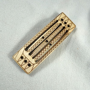 May include: A light-colored wooden cribbage board with etched geometric patterns and three parallel tracks. The board features decorative diamond and triangle shapes, and small holes for pegs.
