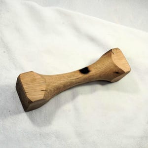 Könnte beinhalten: Eine Holzhantel mit einem dunkelbraunen Brandfleck in der Mitte. Die Hantel ist aus hellem braunem Holz gefertigt und hat an jedem Ende eine quadratische Form.