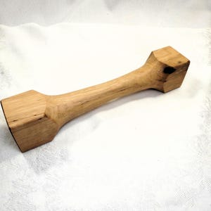 Könnte beinhalten: Ein Holzhammer mit quadratischem Kopf und einem langen, schlanken Stiel. Der Hammer ist aus hellbraunem Holz gefertigt und hat eine natürliche Oberfläche.