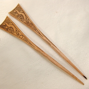 Könnte beinhalten: Zwei Holzhaarnadeln mit aufwendig geschnitzten keltischen Knotenmustern an der Spitze. Die Nadeln sind hellbraun.