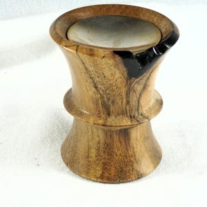 Può includere: Un contenitore cilindrico in legno con parte superiore e inferiore svasate, caratterizzato da una venatura naturale color marrone chiaro e una finitura liscia. L'apertura superiore contiene un inserto circolare metallico e un intarsio in resina nera.