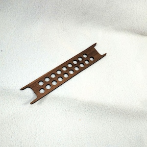 May include: A small, rectangular wooden object with two rows of evenly spaced circular holes. The wood is a rich, dark brown color. The ends of the object have a notched design. This item could be a tool or a decorative piece.