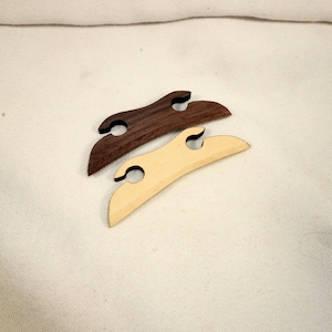 May include: Two wooden shawl pins. One is a dark brown color, and the other is a light tan color. Each pin has a curved shape with two holes and a hook-like design. The pins are displayed on a white fabric background.