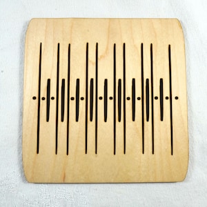 May include: A light-colored wooden coaster with rounded corners. The coaster features a series of vertical black lines and small dots, creating a symmetrical design. The lines vary in length, adding visual interest to the coaster's surface.