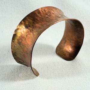 May include: A wide copper cuff bracelet with a hammered finish. The bracelet is made from a single piece of metal and has a slightly curved shape.