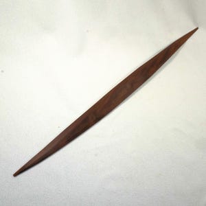 Op de afbeelding: Een lange, slanke houten briefopener met een donkerbruine afwerking. De opener heeft een scherpe, puntige punt en een plat, langwerpig lichaam. De houtnerf is zichtbaar en voegt textuur toe aan het ontwerp. De briefopener is ontworpen voor het openen van enveloppen.