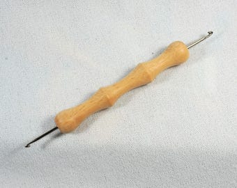 Può includere: Un uncinetto in legno con due uncini in metallo su entrambe le estremità. L'uncinetto è di colore marrone chiaro e ha un manico liscio e arrotondato.