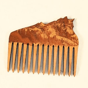 May include: A light brown wooden comb featuring a laser-engraved mountain landscape design on its upper section. The comb has twelve evenly spaced, pointed teeth and a textured, natural wood grain finish across the entire surface area.