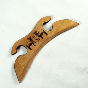 Könnte beinhalten: Flaschenöffner aus Holz mit geschwungenem Design und zwei Aussparungen. Der Öffner zeigt zwei gravierte Schafillustrationen. Das Holz hat eine hellbraune Farbe mit glatter Oberfläche. Der Öffner ist zum Öffnen von Flaschen bestimmt.