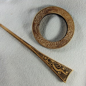 Könnte beinhalten: Ein Holzhaarspangen mit komplizierten keltischen Knotenmustern. Die Spange ist lang und schlank mit einem spitzen Ende. Die Spange wird von einer runden Holzbrosche mit einem ähnlichen keltischen Knotenmuster begleitet.