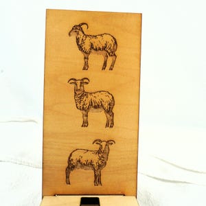 Könnte beinhalten: Ein Holzschild mit drei lasergravierten Bildern von Schafen. Die Schafe sind nach rechts gerichtet und haben lockige Hörner.