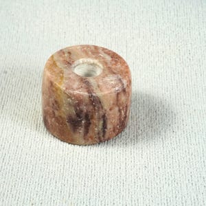 May include: A cylindrical stone object with a polished, marbled surface featuring swirling patterns of reddish-brown, tan, and cream colors. The object has a circular opening in the center of its top surface, creating a hollow, bead-like structure.