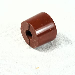 May include: A cylindrical brown bead with a smooth, glossy finish and a central hole. The bead has a flat circular face with a small notch extending from the center hole to the outer edge, showing a matte texture on the interior surface.