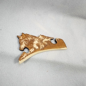 May include: A wooden puzzle piece with a wolf and mountain design. The wolf is detailed with textured fur, and mountains are in the background. The piece is light brown with a cream-coloured edge.