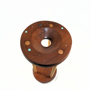 May include: A handcrafted wooden sculpture with a dark brown finish. The top features a circular design with a central hole and inlaid decorative elements in various colors. The sculpture is supported by three wooden legs.