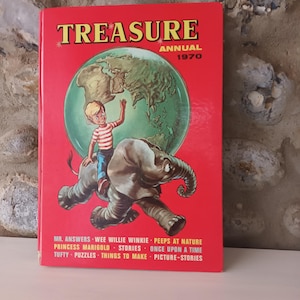 Puede incluir: Un libro vintage "Treasure Annual 1970" con una cubierta roja. La portada muestra a un niño montado en un elefante sobre un mapa del mundo. El título está en letras blancas. El libro contiene historias.