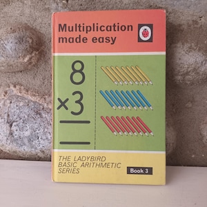 Könnte beinhalten: Ein Vintage-Buch der Ladybird Basic Arithmetic Series, Buch 3, mit dem Titel "Multiplication made easy". Das Cover ist orange und grün, mit einer Multiplikationsaufgabe und Bleistiftillustrationen. Das Buch liegt auf einer weißen Oberfläche.