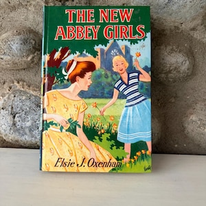 Puede incluir: Un libro vintage titulado "The New Abbey Girls" con una cubierta verde. La portada muestra a dos mujeres con vestidos, una amarilla y otra azul, en un jardín. El nombre de la autora, Elsie J. Oxenham, está impreso en la portada.