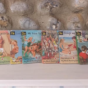 Op de afbeelding: Een verzameling vintage Ladybird Key Words Reading Scheme boeken. De covers tonen kleurrijke illustraties van spelende kinderen, met titels als "Play with us" en "We have fun". De boeken zijn gerangschikt op een wit oppervlak.
