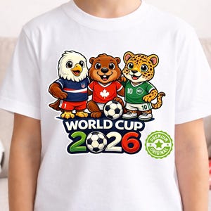 May include: White t-shirt with three cartoon mascots in football jerseys: an eagle, a beaver, and a jaguar, with a football. The shirt displays "WORLD CUP 2026" and a "Premium Original" badge.