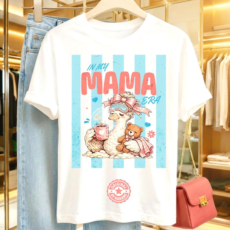 May include: White t-shirt with a graphic featuring a cartoon duck wearing a bow, eye mask, and holding a mug. The text "IN MY MAMA ERA" is displayed in pink and blue. A small teddy bear is also featured.
