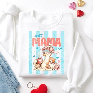 May include: White sweatshirt with a graphic featuring a duck wearing a sleep mask and holding a mug and a teddy bear. The text "IN MY MAMA ERA" is displayed in pink and blue. The design is set against a backdrop of blue stripes.