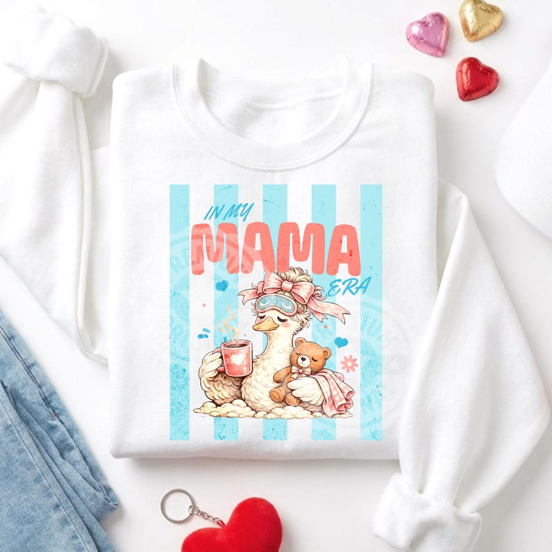 May include: White sweatshirt with a graphic featuring a duck wearing a sleep mask and holding a mug and a teddy bear. The text "IN MY MAMA ERA" is displayed in pink and blue. The design is set against a backdrop of blue stripes.