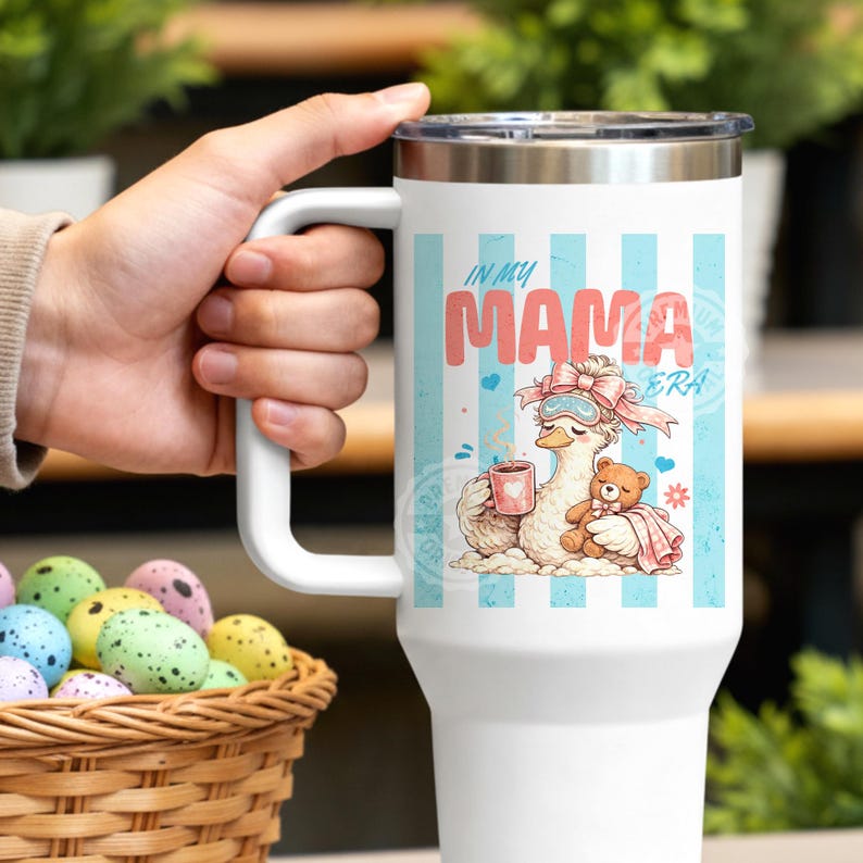 May include: White travel mug with a handle, featuring a cartoon llama wearing a sleep mask and holding a mug. The text "IN MY MAMA ERA" is displayed on a light blue striped background. A teddy bear is also in the image.