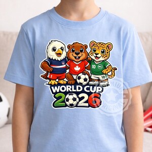 May include: Pale blue t-shirt with a graphic of three cartoon mascots: an eagle, a beaver, and a jaguar, in football kits. The shirt displays "WORLD CUP 2026" with footballs. The eagle mascot wears a USA kit.