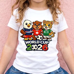 May include: White t-shirt with cartoon mascots of the USA, Canada, and Mexico, each in a jersey and holding a football. The shirt has the text "WORLD CUP 2026" in green, black, and red. The shirt is worn by a child.