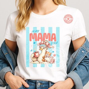 May include: White t-shirt with a graphic of a cartoon goose wearing a sleep mask and holding a mug, with a teddy bear. The text "IN MY MAMA ERA" is above the goose, with blue stripes in the background.