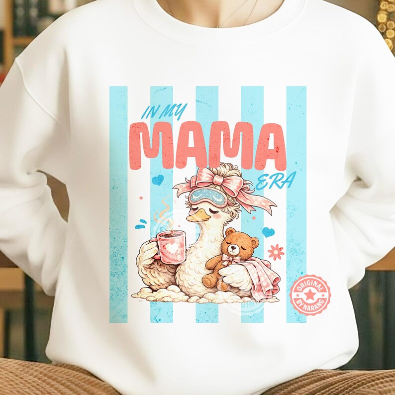 May include: White sweatshirt with a graphic featuring a cartoon goose wearing a sleep mask and holding a mug and teddy bear. The text "IN MY MAMA ERA" is displayed above the goose, with blue and white striped background.