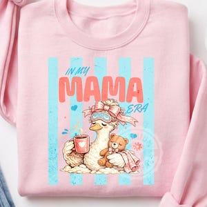 May include: Pink sweatshirt with a graphic featuring a cartoon duck wearing a sleep mask and holding a mug. The text "IN MY MAMA ERA" is displayed in coral and teal. A teddy bear is nestled next to the duck.