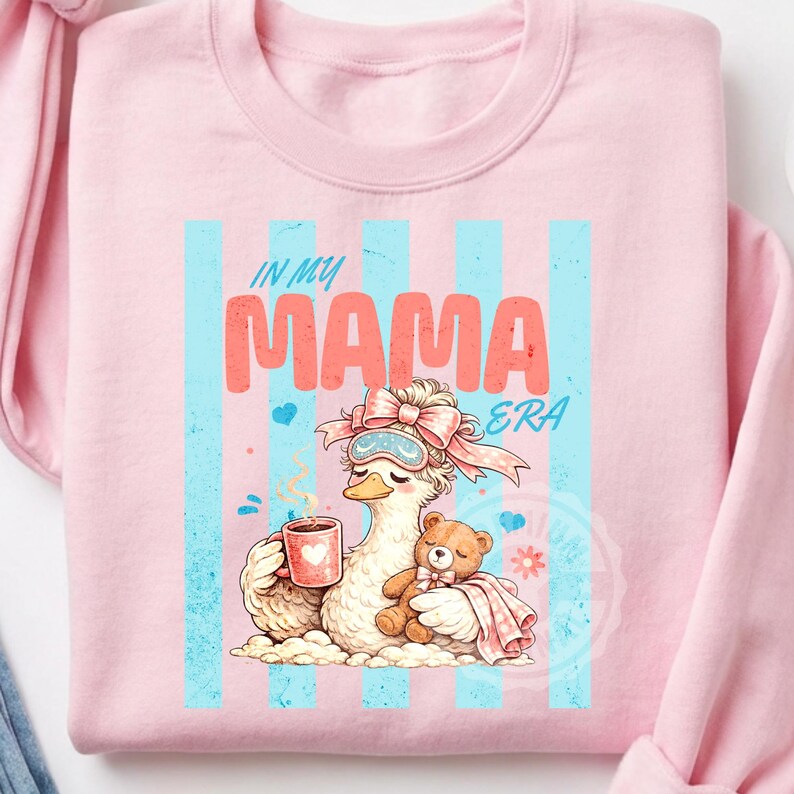 May include: Pink sweatshirt with a graphic featuring a cartoon duck wearing a sleep mask and holding a mug. The text "IN MY MAMA ERA" is displayed in coral and teal. A teddy bear is nestled next to the duck.