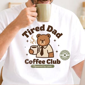 Puede incluir: Camiseta blanca con un oso pardo sosteniendo una taza de café, con el texto "Tired Dad Coffee Club" y "Powered by Love". El oso lleva camisa y corbata. También se ve una insignia "Premium Original".