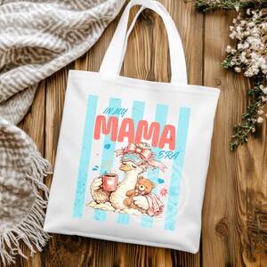 May include: White tote bag with a graphic of a cartoon goose wearing a sleep mask and holding a mug, with a teddy bear. The text "IN MY MAMA ERA" is displayed in pink and blue.