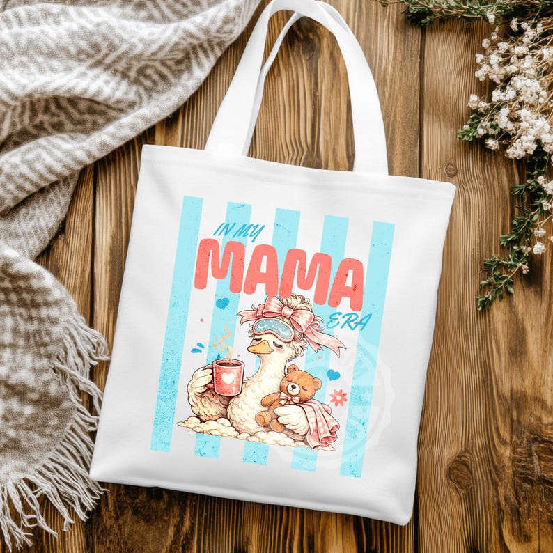 May include: White tote bag with a graphic of a cartoon goose wearing a sleep mask and holding a mug, with a teddy bear. The text "IN MY MAMA ERA" is displayed in pink and blue.