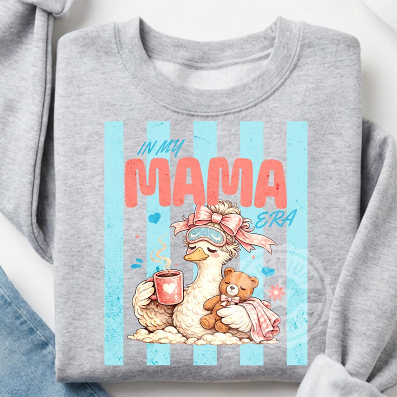 May include: A light gray sweatshirt with the text "IN MY MAMA ERA" in pink and blue. The graphic features a cartoon duck wearing a sleep mask and holding a mug and teddy bear. Blue vertical stripes are behind the graphic.