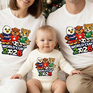 May include: White t-shirts and a baby onesie with a cartoon design of an eagle, a bear, and a leopard in soccer kits. The text "WORLD CUP 2026" is printed below the characters, with a football graphic.
