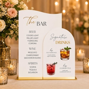 May include: A white acrylic bar menu sign with gold accents featuring The Bar list of beer, wine, and spirits, and a Signature Drinks section with images of an Old Fashioned and a Cranberry Mule, all mounted on a gold metal base.