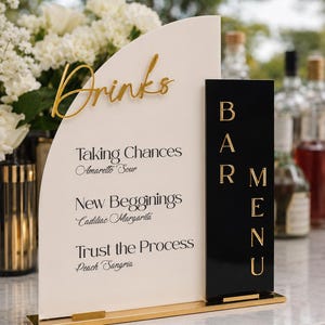 May include: A semi-circular cream acrylic sign with gold lettering reading Drinks, Taking Chances Amaretto Sour, New Begginings Cadillac Margarita, and Trust the Process Peach Sangria, attached to a black rectangular Bar Menu sign on a gold base.