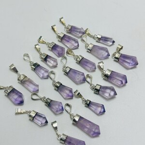 Könnte beinhalten: Eine Sammlung von Amethyst-Kristallanhängern mit silberfarbenen Metallfassungen. Die Anhänger sind helllila und facettiert. Sie sind auf einer weißen Oberfläche angeordnet.